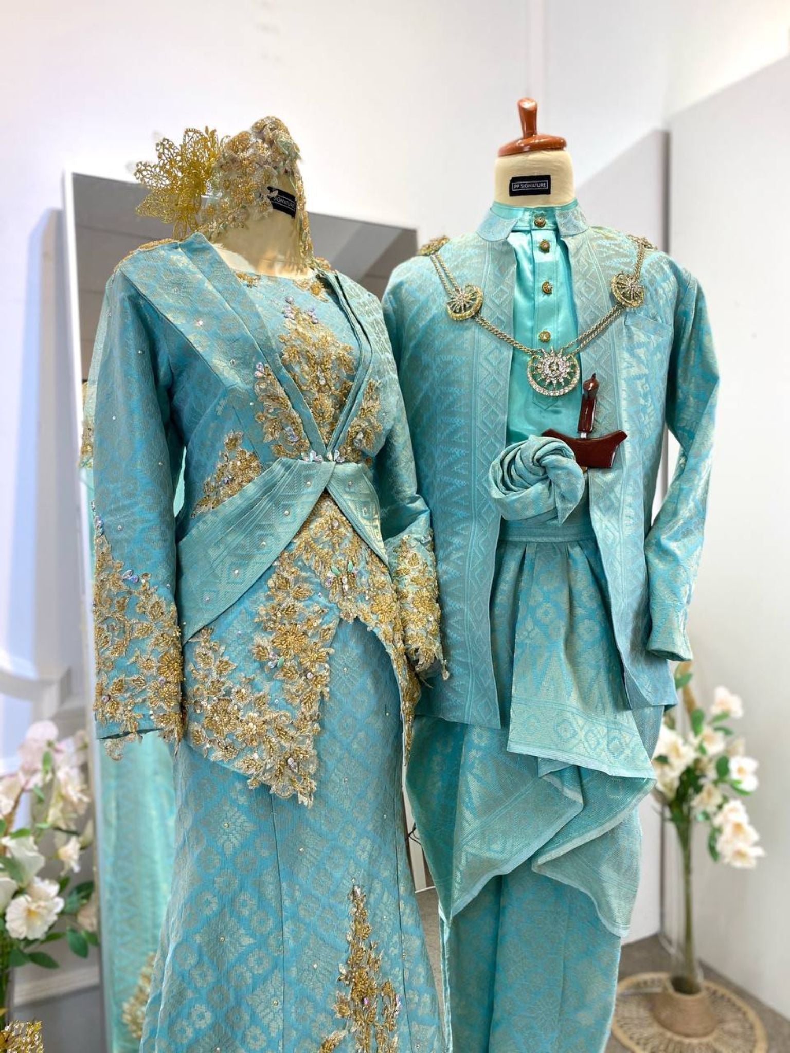 The SURAYA Malay wedding dress features a two-piece teal green songket dress with light gold accents, perfect for the traditional Baju Sanding ceremony. The bride's ensemble is complemented by the groom's three-piece Kot Raihan attire.