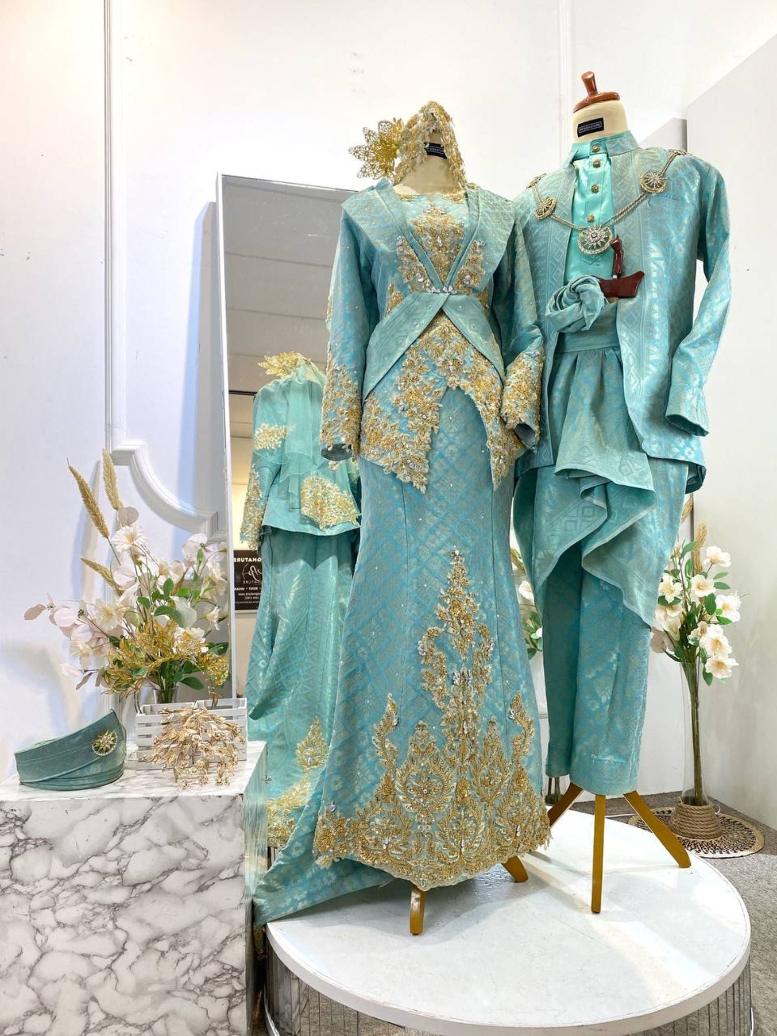 The SURAYA Malay wedding dress features a two-piece teal green songket dress with light gold accents, perfect for the traditional Baju Sanding ceremony. The bride's ensemble is complemented by the groom's three-piece Kot Raihan attire.